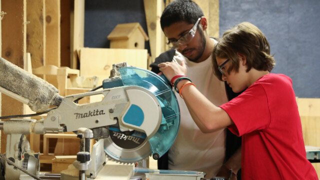 A Kingsley Pines counselor guides a camper using a saw in woodworking